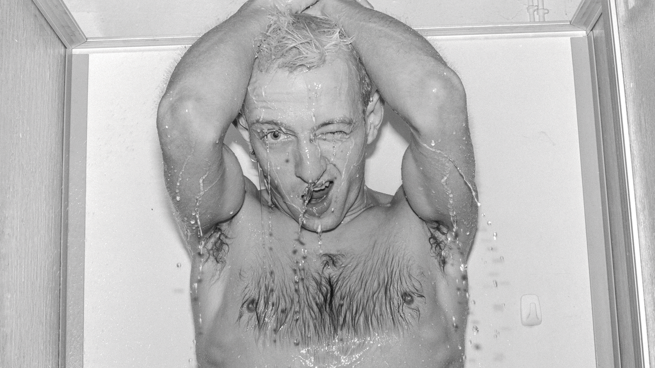 Porträt von Jürgen Baldiga in schwarz-weiß. Von der Brust aufwärts unter fließendem Wasser fotografiert, zwinkert Baldiga in die Kamera.
