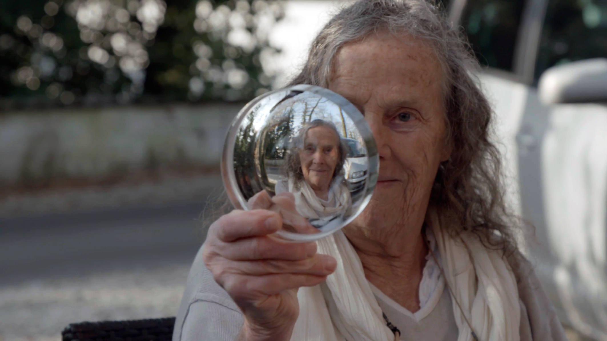 Porträt von Mary Bauermeister. Mary blickt durch ein Glasobjekt in die Kamera, durch das wir ihr Gesicht scharf sehen, und im Hintergrund können wir sie unscharf sehen.
